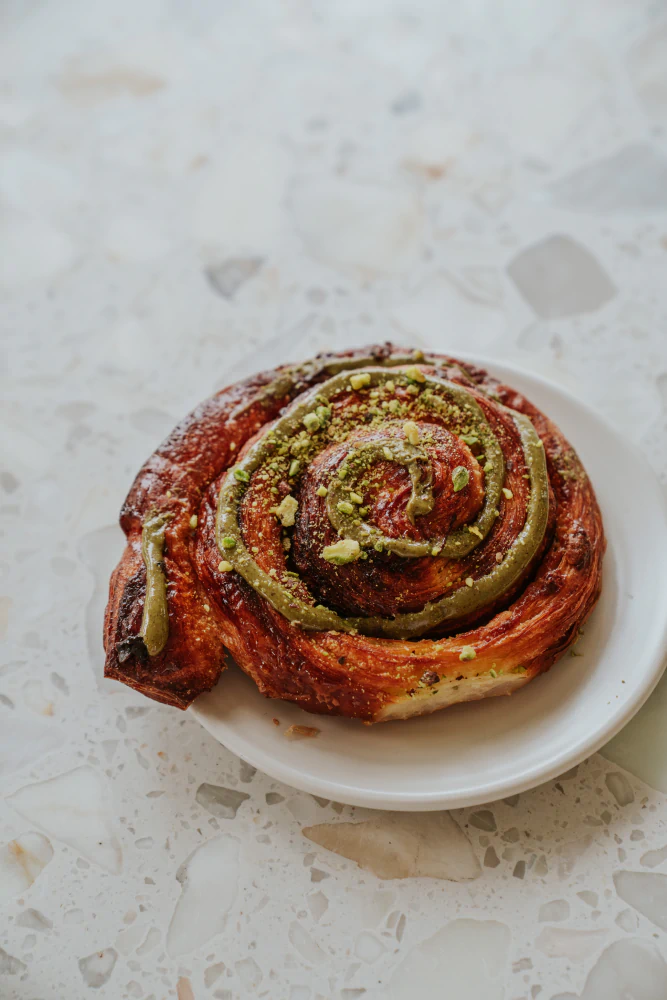 Close-up of a spiral pastry topped with green pistachio cream and crushed pistachios on a white plate.