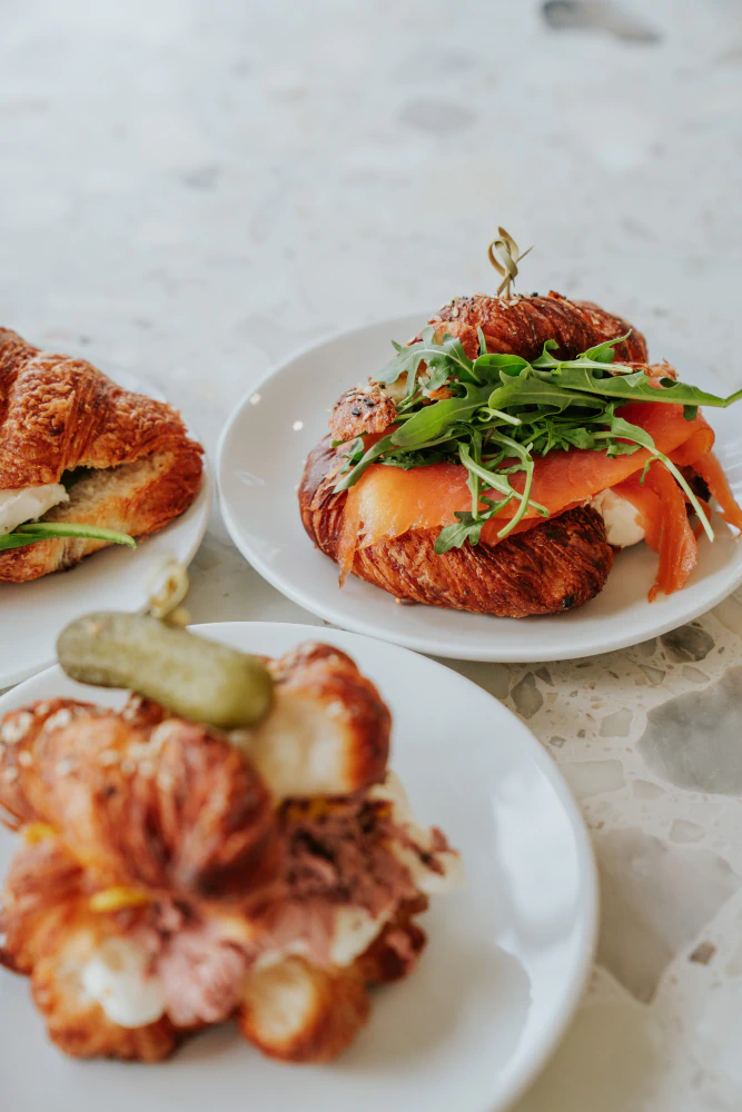 Three croissant sandwiches with fillings including smoked salmon and arugula, a small pickle on one sandwich, and cream cheese.