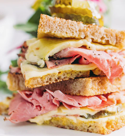 Close-up of a stacked sandwich with roast beef, melted cheese, pickles, and whole grain bread.