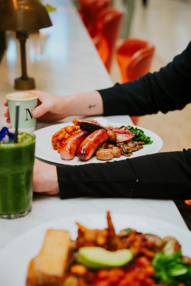 Person holding a cup next to a plate with sausages, baked beans, mushrooms, greens, and other cooked breakfast items on a counter.