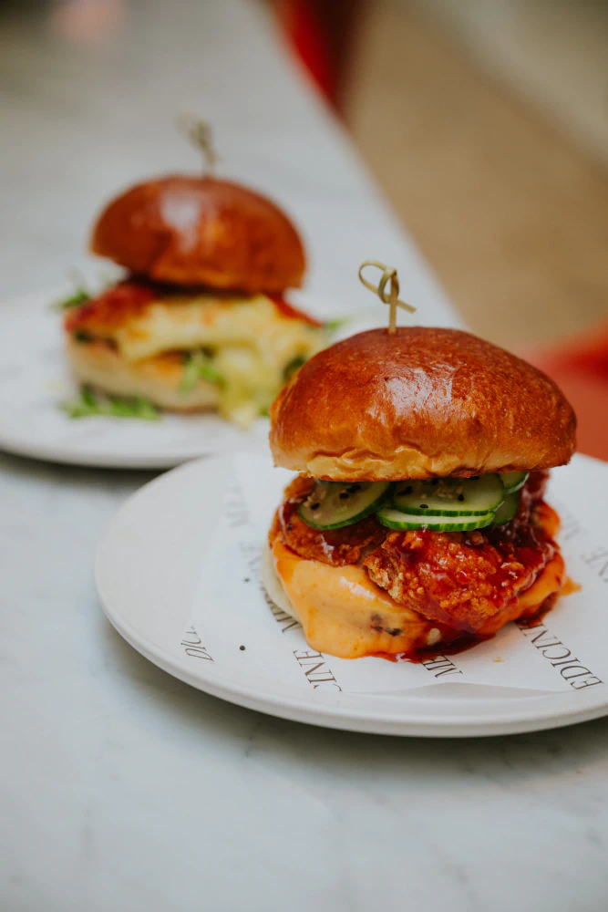 Close-up of a spicy fried chicken sandwich with cucumber slices and sauce on a brioche bun served on a white plate.