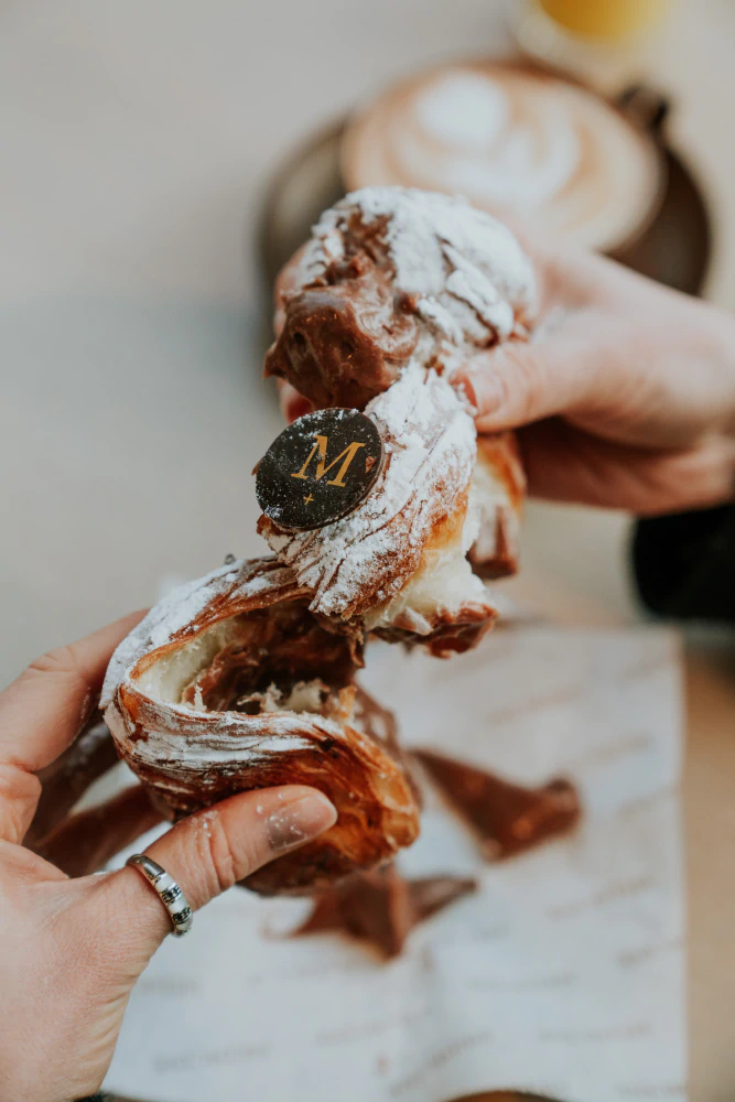 Hands tearing apart a powdered sugar-coated chocolate croissant with a dark chocolate piece marked with a gold 'M'.