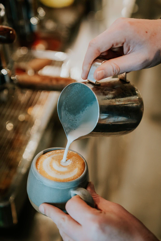 Hands pouring steamed milk from a metal pitcher into a blue mug of coffee to create latte art.