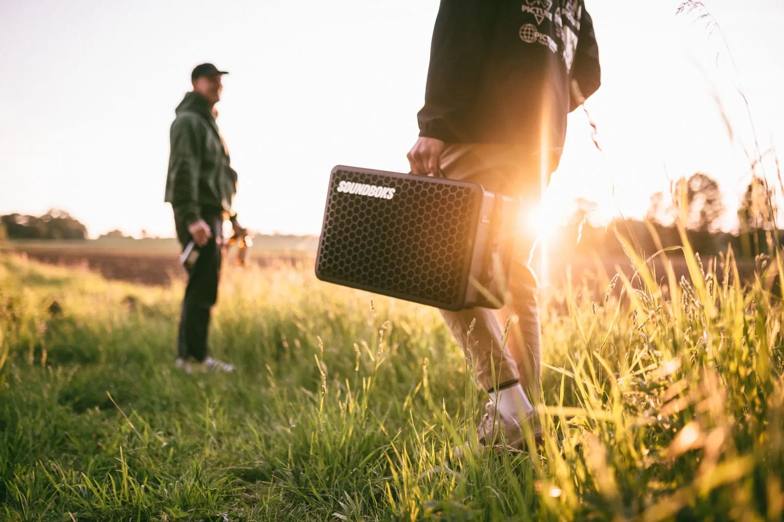 Zwei Männer im Freien, einer trägt einen tragbaren SOUNDBOKS-Lautsprecher bei Sonnenuntergang. Foto/Video von Felix Weise, Medienproduktion für Sport, Outdoor & Events im Allgäu, Kempten und Bayern.