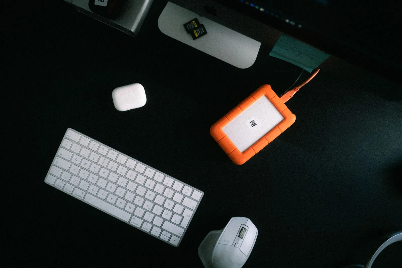 Tastatur, Maus, weiße kabellose Ohrhörer, Speicherkarten und orange externe Festplatte auf schwarzem Tisch. Foto/Video von Felix Weise, Medienproduktion für Sport, Outdoor & Events im Allgäu, Kempten und Bayern.