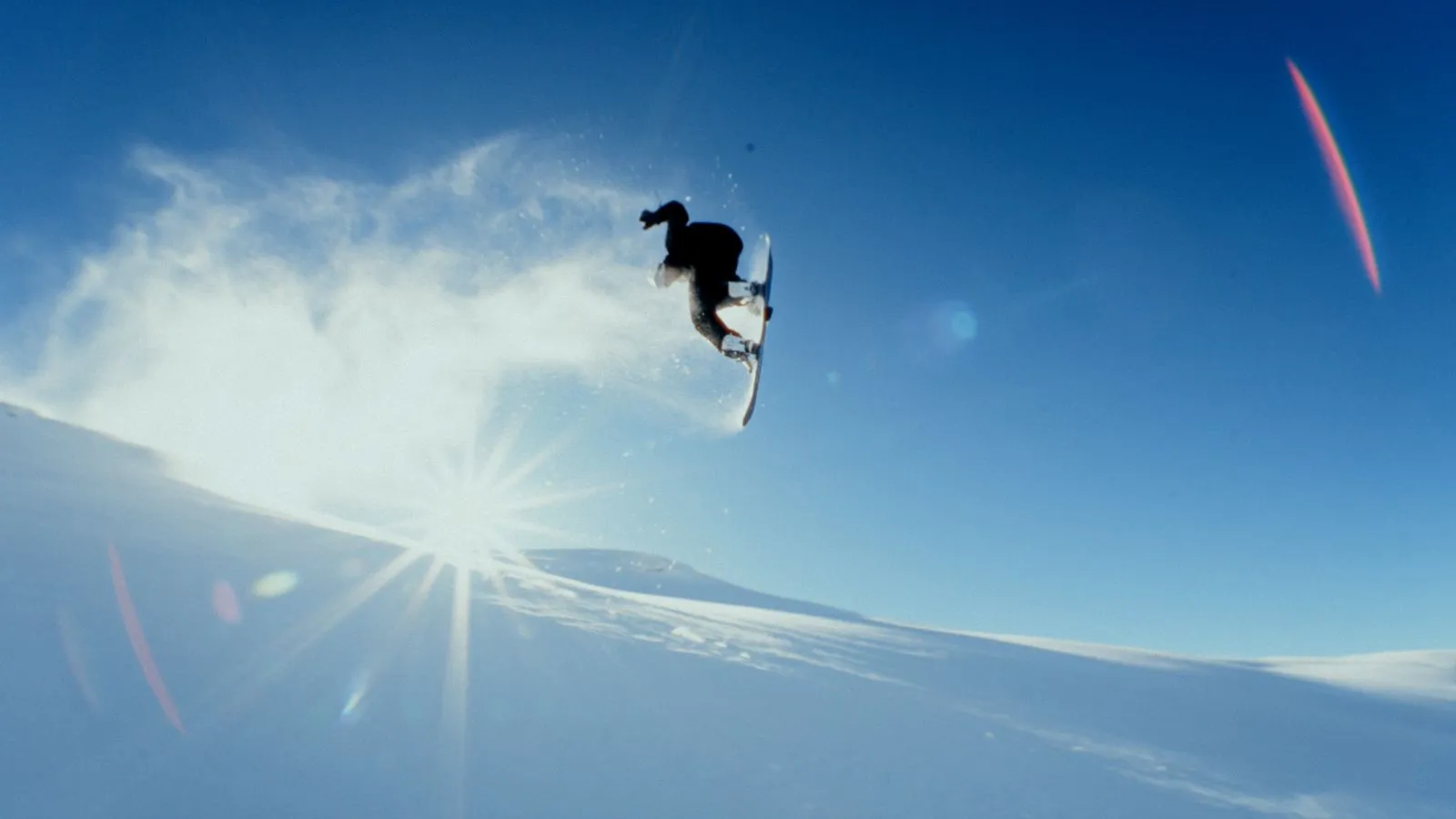 Snowboarder führt einen Sprung unter klarem blauem Himmel mit Sonnenlicht und Schneestaub aus. Foto/Video von Felix Weise, Medienproduktion für Sport, Outdoor & Events im Allgäu, Kempten und Bayern.