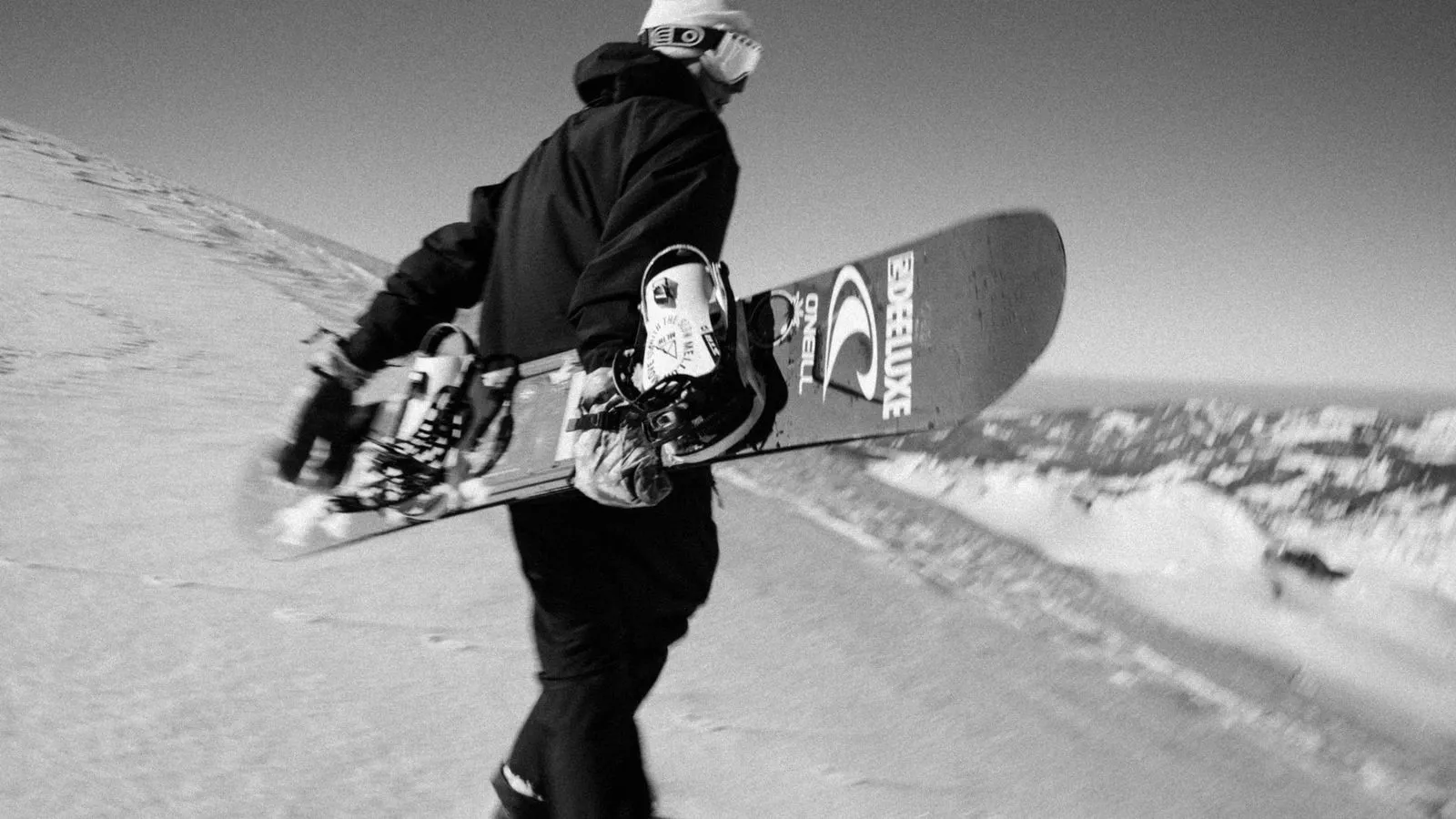 Snowboarder in Winterkleidung trägt Snowboard auf verschneitem Berg mit Bergpanorama im Hintergrund. Foto/Video von Felix Weise, Medienproduktion für Sport, Outdoor & Events im Allgäu, Kempten und Bayern.