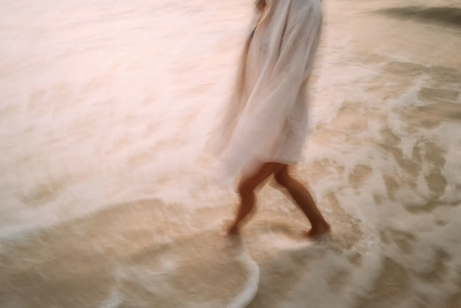 Unscharfe Aufnahme von einer Person im weißen Kleid, die am Strand im Wasser läuft. Foto/Video von Felix Weise, Medienproduktion für Sport, Outdoor & Events im Allgäu, Kempten und Bayern.