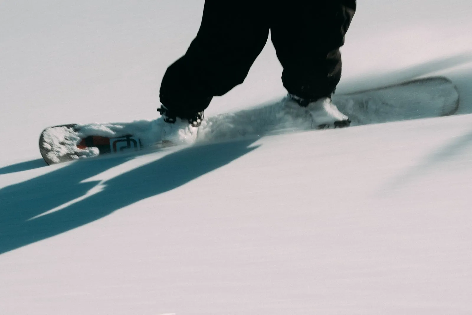 Person snowboarding on fresh snow wearing black pants and white boots, casting a shadow. Foto/Video von Felix Weise, Medienproduktion für Sport, Outdoor & Events im Allgäu, Kempten und Bayern.
