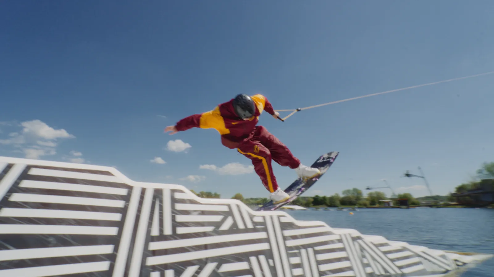 Wakeboarder in rotem und gelbem Anzug springt über Rampe auf einem See. Foto/Video von Felix Weise, Medienproduktion für Sport, Outdoor & Events im Allgäu, Kempten und Bayern.