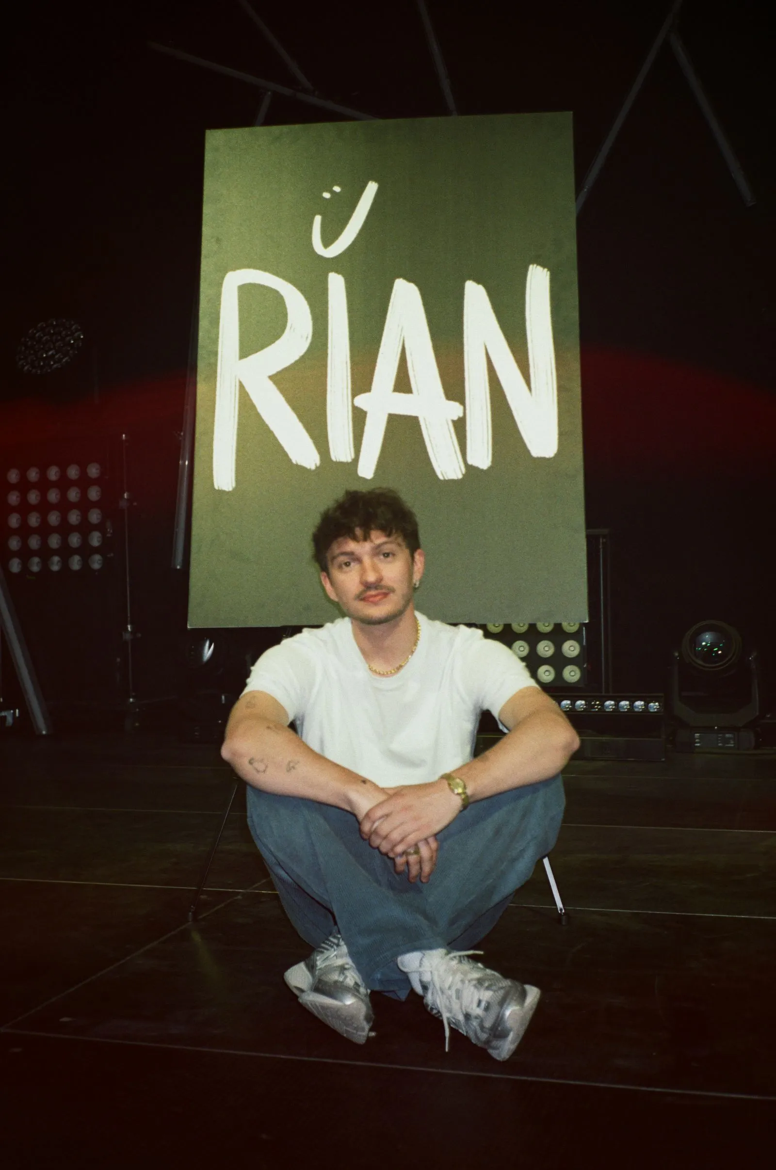 Der Künstler RIAN sitzt auf dem Boden vor einem Schild mit der Aufschrift „RIAN“ in großen Buchstaben. Foto/Video von Felix Weise, Medienproduktion für Sport, Outdoor & Events im Allgäu, Kempten und Bayern.