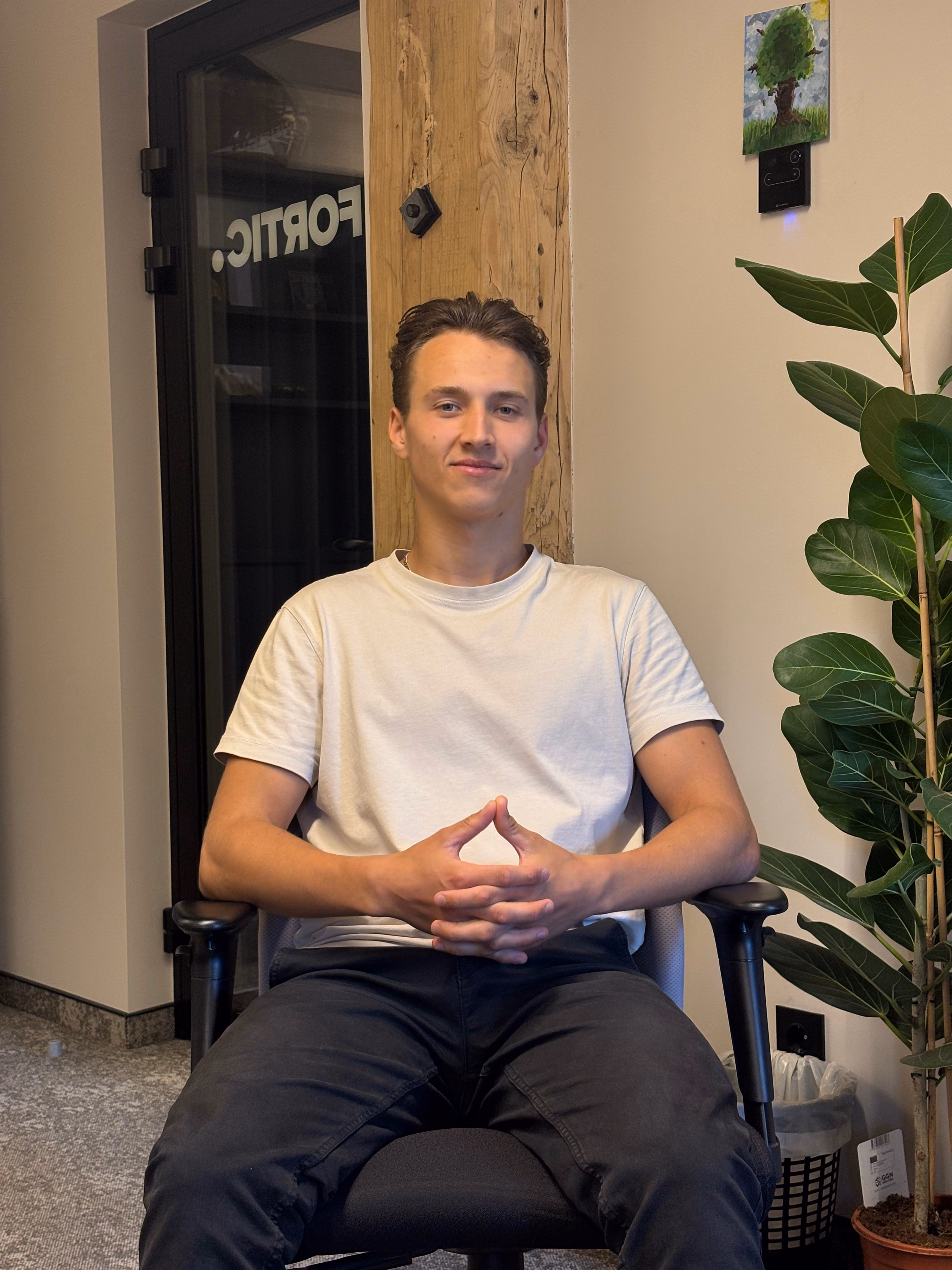 Young man with short brown hair wearing a white t-shirt and black pants sitting on a black office chair in an indoor setting, with a wooden beam, potted plant, trash can, and door with partially visible text behind him.