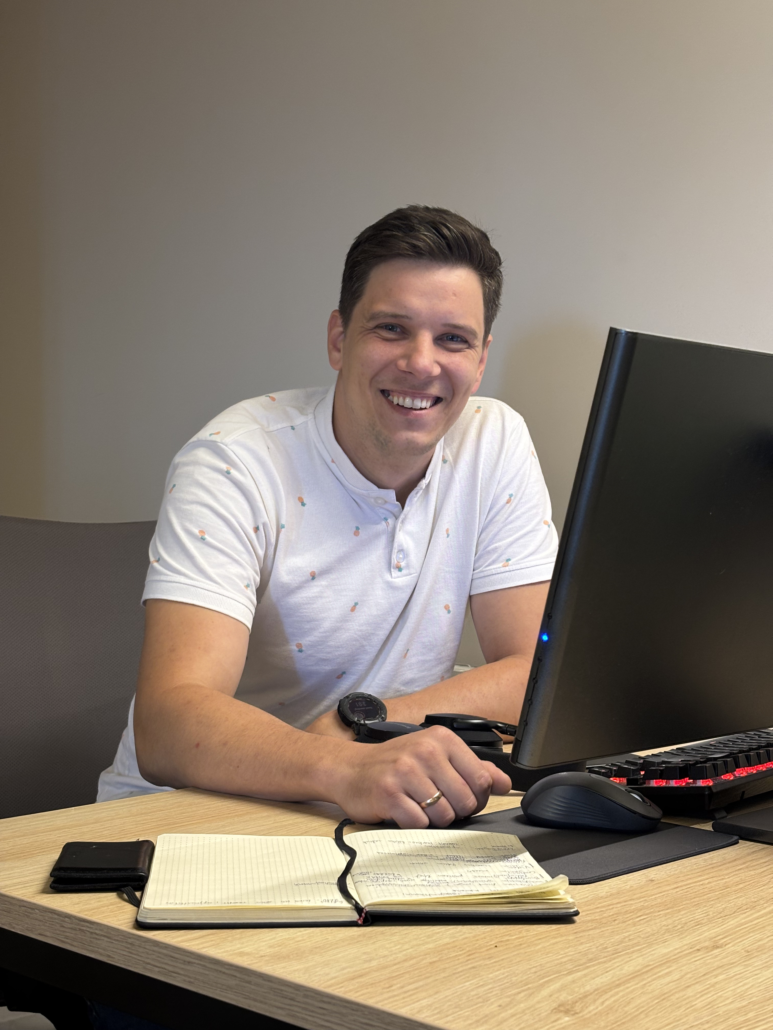 Smiling man with short brown hair wearing a white shirt with small orange and green patterns, sitting at a desk with a computer, keyboard, mouse, open notebook, and wallet.