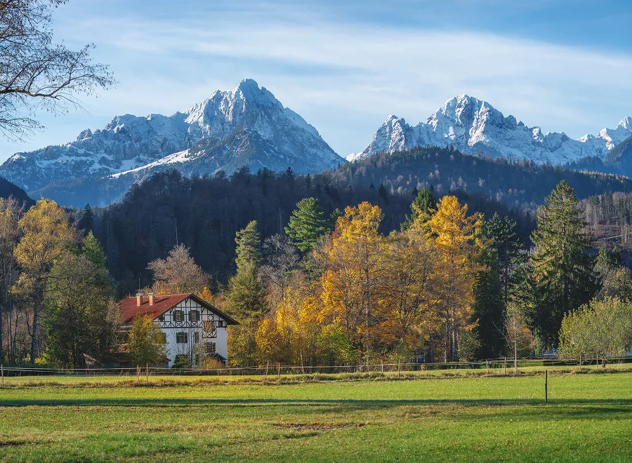 Photo of a house in a green meadow with trees with visible mountains in the background