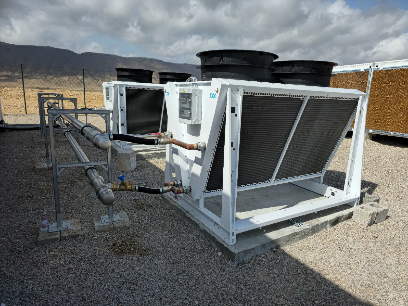 Two industrial cooling units with black fans and connected metal pipes on gravel under a cloudy sky.