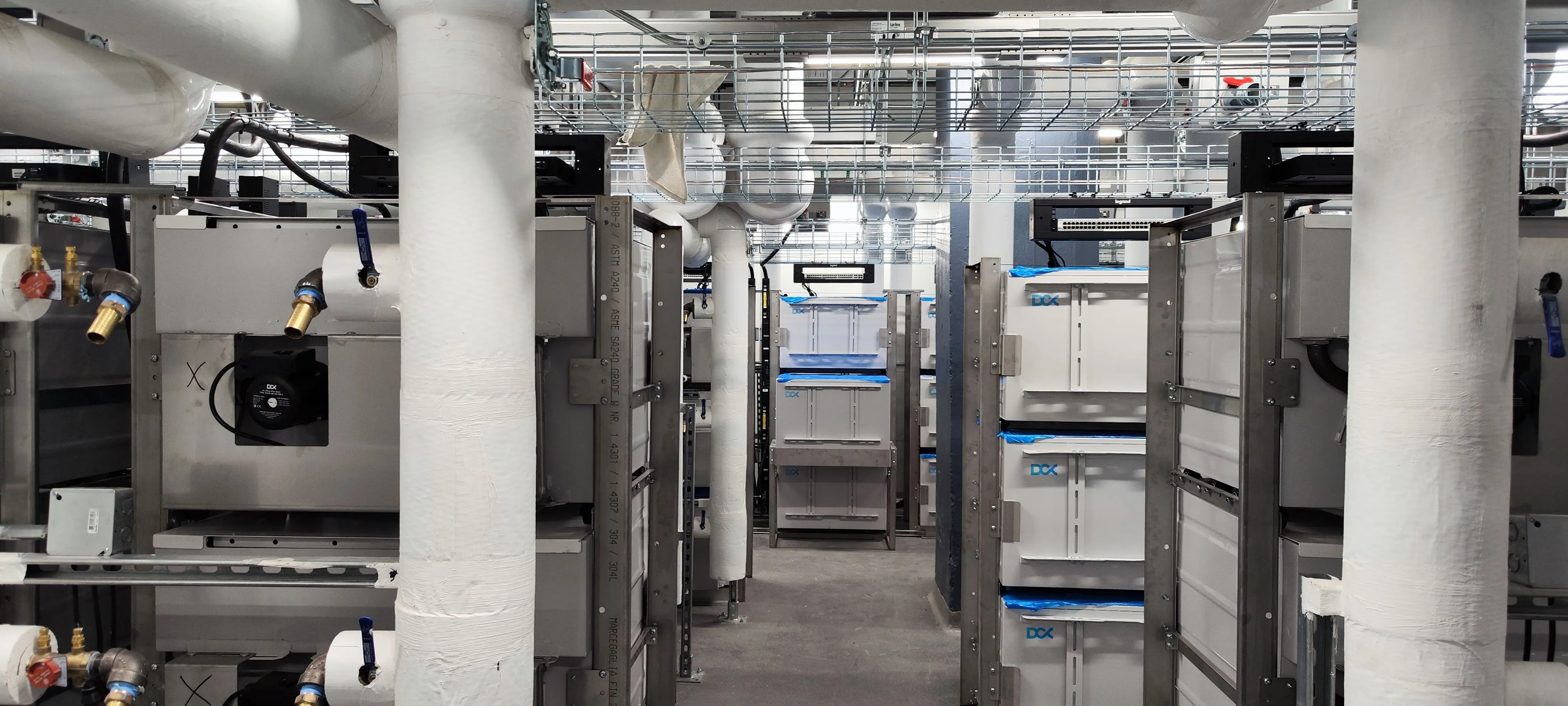 Industrial room with insulated pipes and stacked white metal HVAC units and equipment.