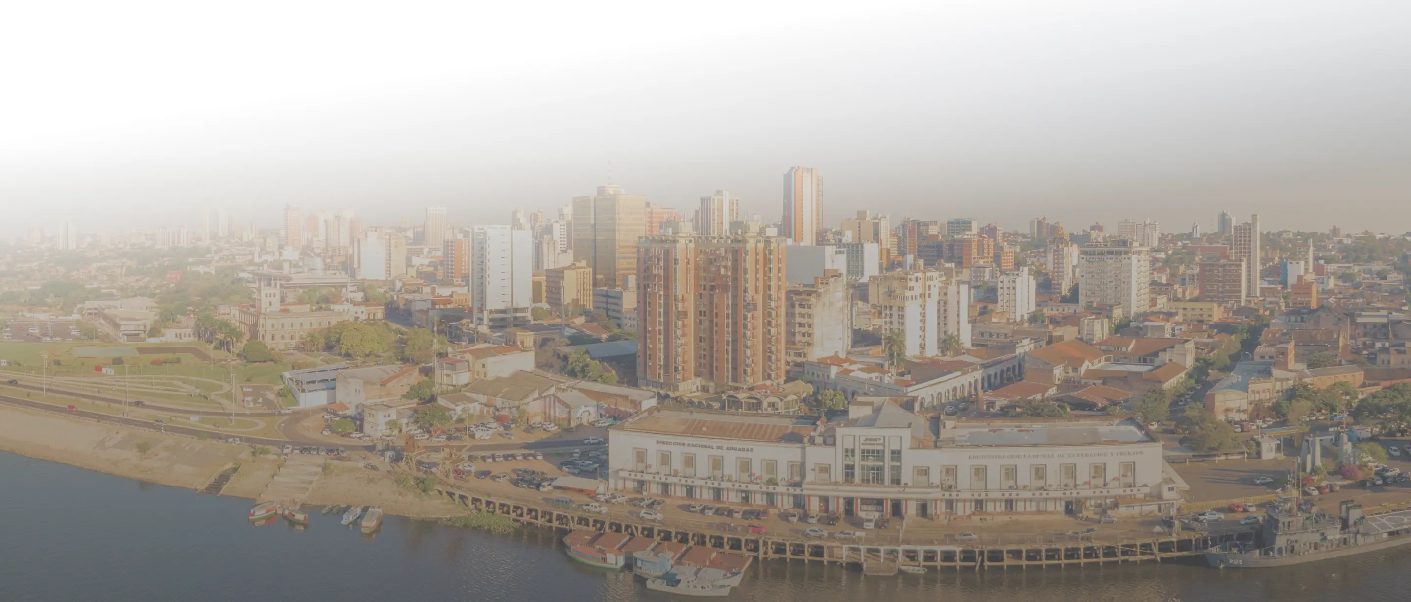 Cityscape showing mixed buildings and boats docked along a waterfront pier under hazy sky.