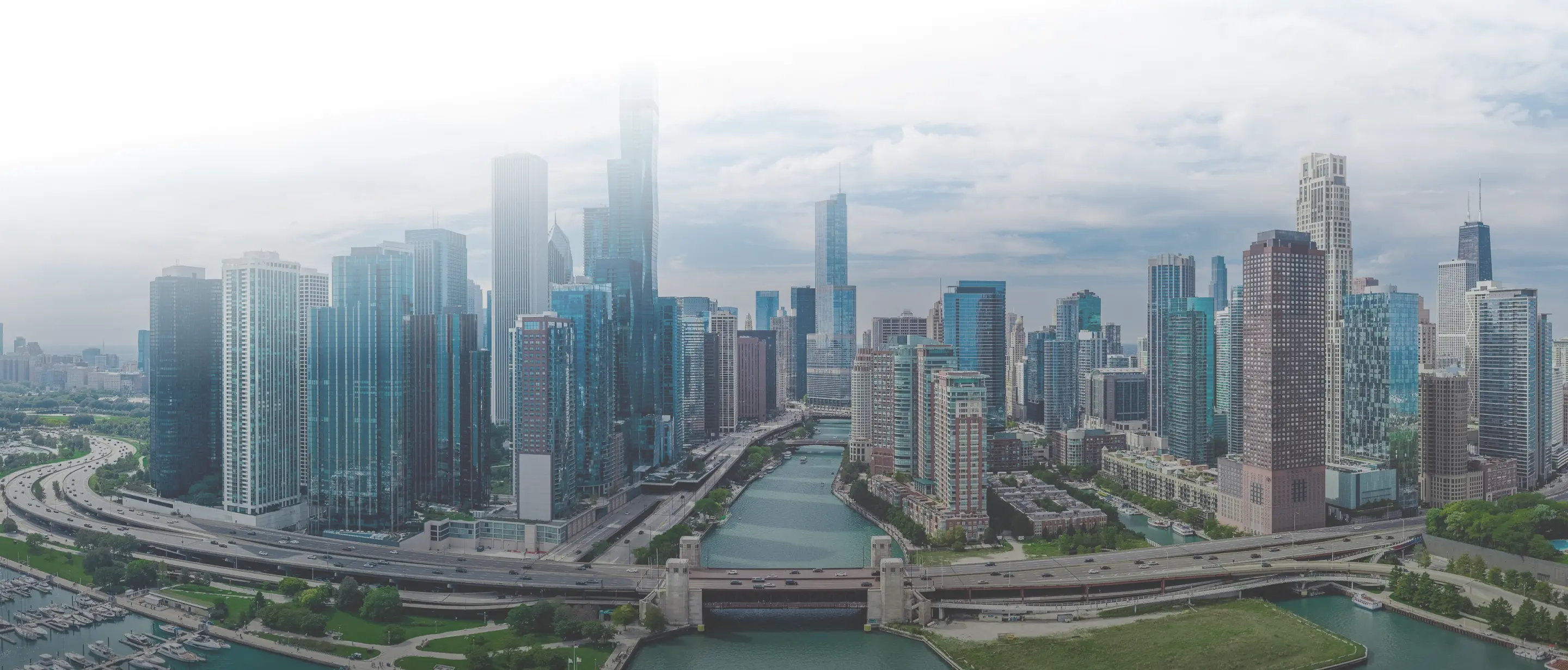 Aerial view of Chicago skyline with river, bridges, highways, and high-rise buildings under cloudy sky.