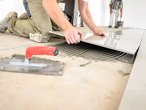 Person verlegt glatte weiße Bodenfliesen auf Kleber mit einer Kelle auf dem Boden.