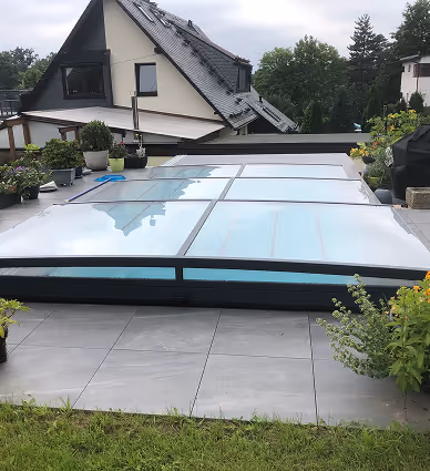 Modernes, flach abgedecktes Schwimmbad auf einer Terrasse mit grauen Fliesen, umgeben von Töpfen mit Pflanzen und Blick auf ein benachbartes Haus mit schwarzem Dach.