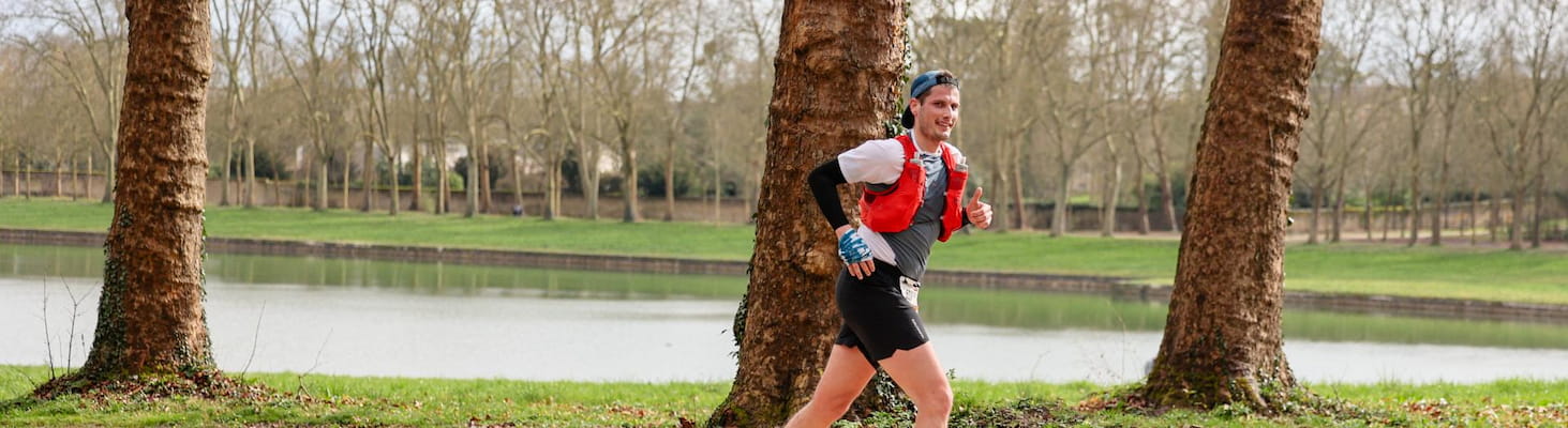 Un coureur souriant en plein effort sur un sentier bordé d’arbres, longeant un plan d’eau en pleine nature lors de l’EcoTrail Paris.