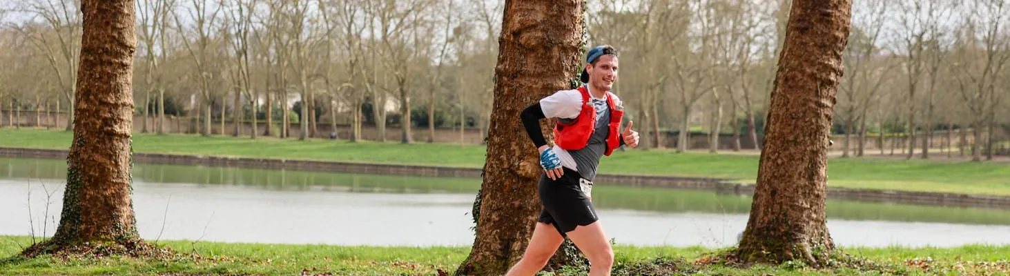 Un coureur souriant en plein effort sur un sentier bordé d’arbres, longeant un plan d’eau en pleine nature lors de l’EcoTrail Paris.