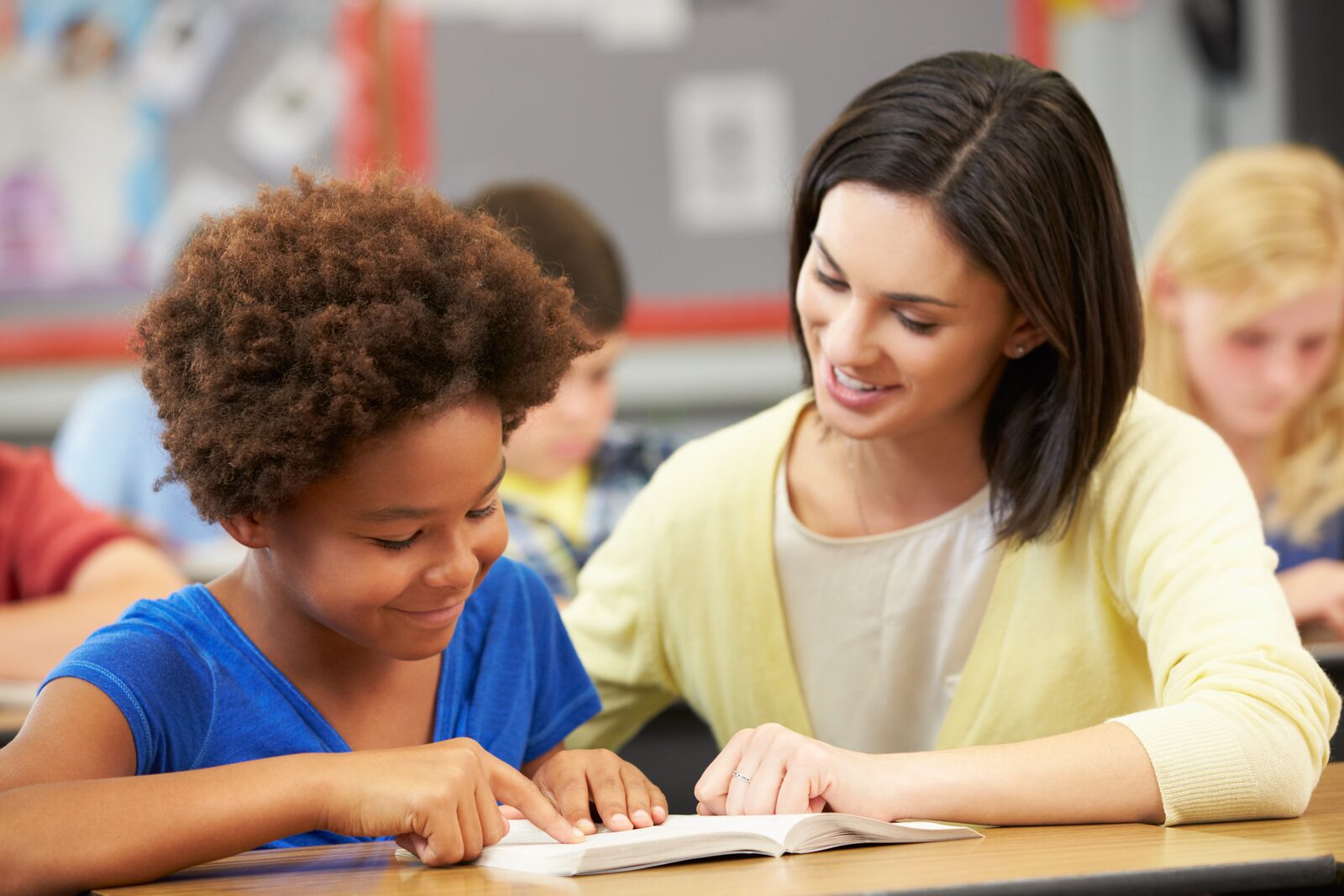 A teacher leans in to read a book with a student in class.