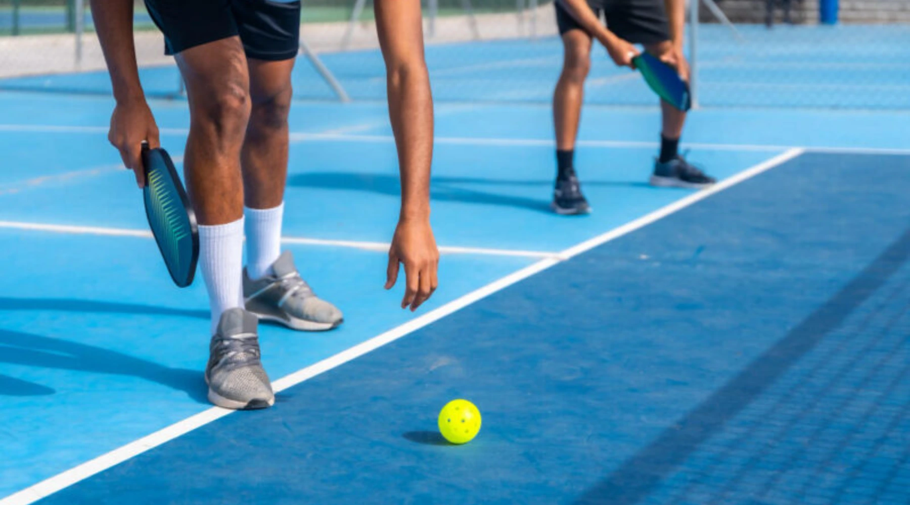 Two men on a pickleball court, one bending to pick up a bright yellow ball with paddle ready