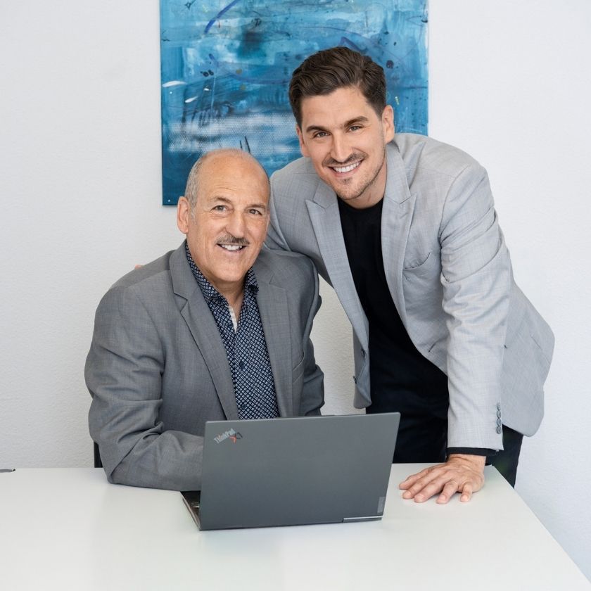Yves und Urs Pfeuti von YU Broker AG lächeln in die Kamera, einer sitzt vor einem geöffneten Laptop.