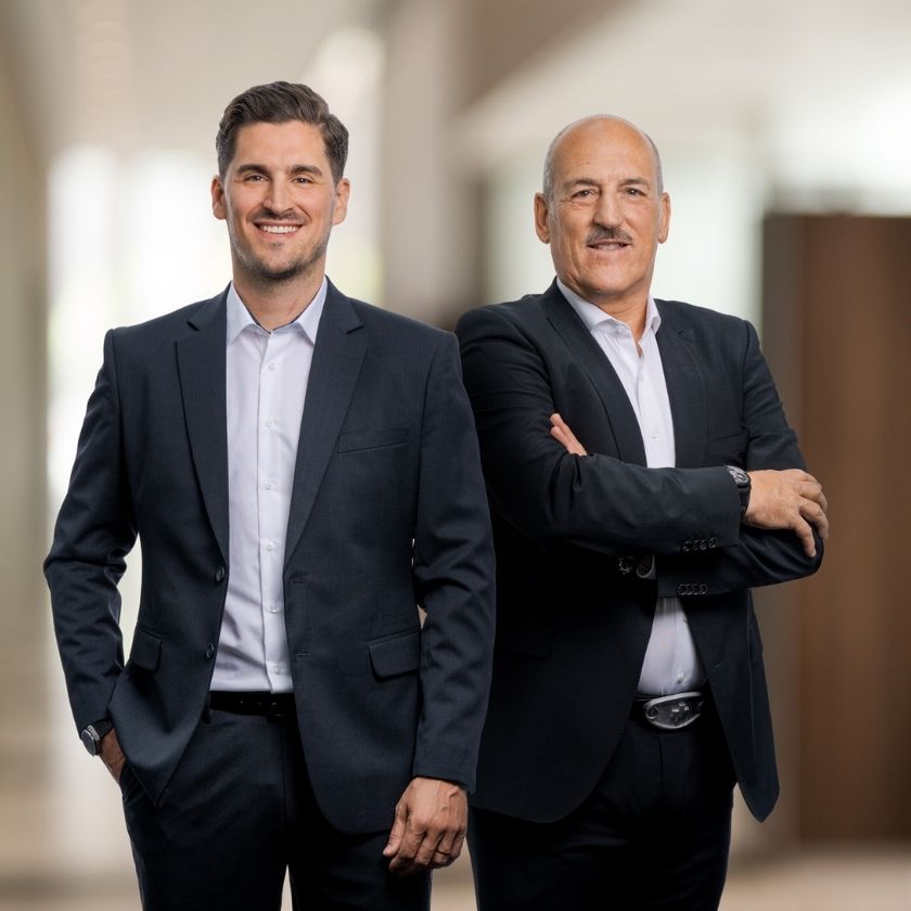 Yves und Urs Pfeuti von YU Broker AG in dunklen Anzügen stehen nebeneinander in einem modernen, unscharf dargestellten Innenraum