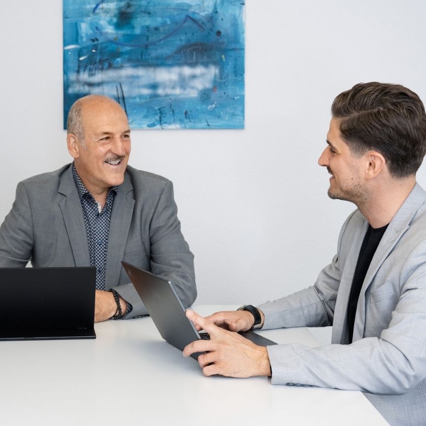 Yves und Urs Pfeuti von YU Broker AG in grauen Sakkos sitzen sich an einem Tisch mit Laptops gegenüber und unterhalten sich freundlich