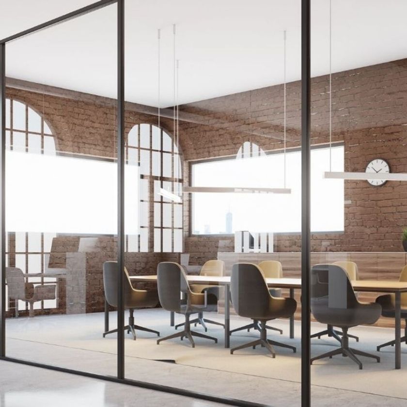 Moderner Besprechungsraum mit Glaswänden, Backsteinmauern, großem Konferenztisch und mehreren Bürostühlen; große Fenster lassen Tageslicht herein, eine Uhr hängt an der Wand.