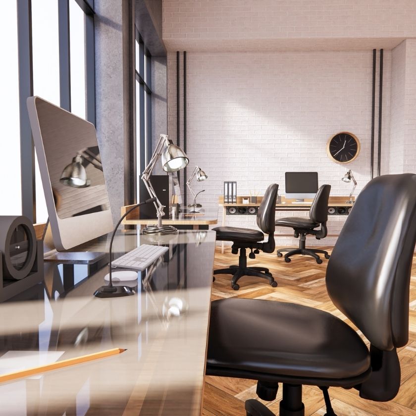 Modern eingerichtetes Großraumbüro mit Schreibtischen, Bürostühlen, iMac-Computern und Designerlampen vor einer weißen Backsteinwand; eine Wanduhr und große Fenster sorgen für ein helles, stilvolles Arbeitsumfeld.