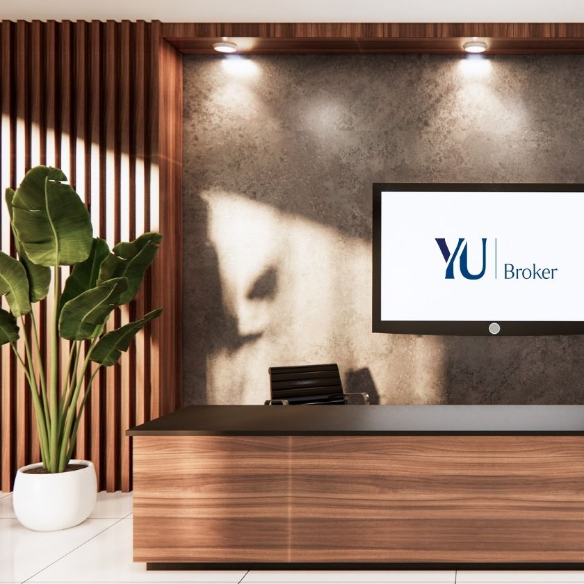 Moderner Empfangsbereich mit Holztheke und vertäfelter Wand, daneben eine große grüne Pflanze in weißem Topf; an der Wand hängt ein Bildschirm mit dem Logo „YU Broker“, stilvoll beleuchtet durch drei Deckenstrahler.