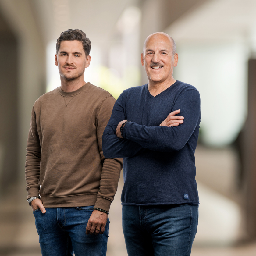 Yves und Urs Pfeuti von YU Broker AG in Worb in Freizeitkleidung stehen nebeneinander in einem modernen, unscharf dargestellten Innenraum