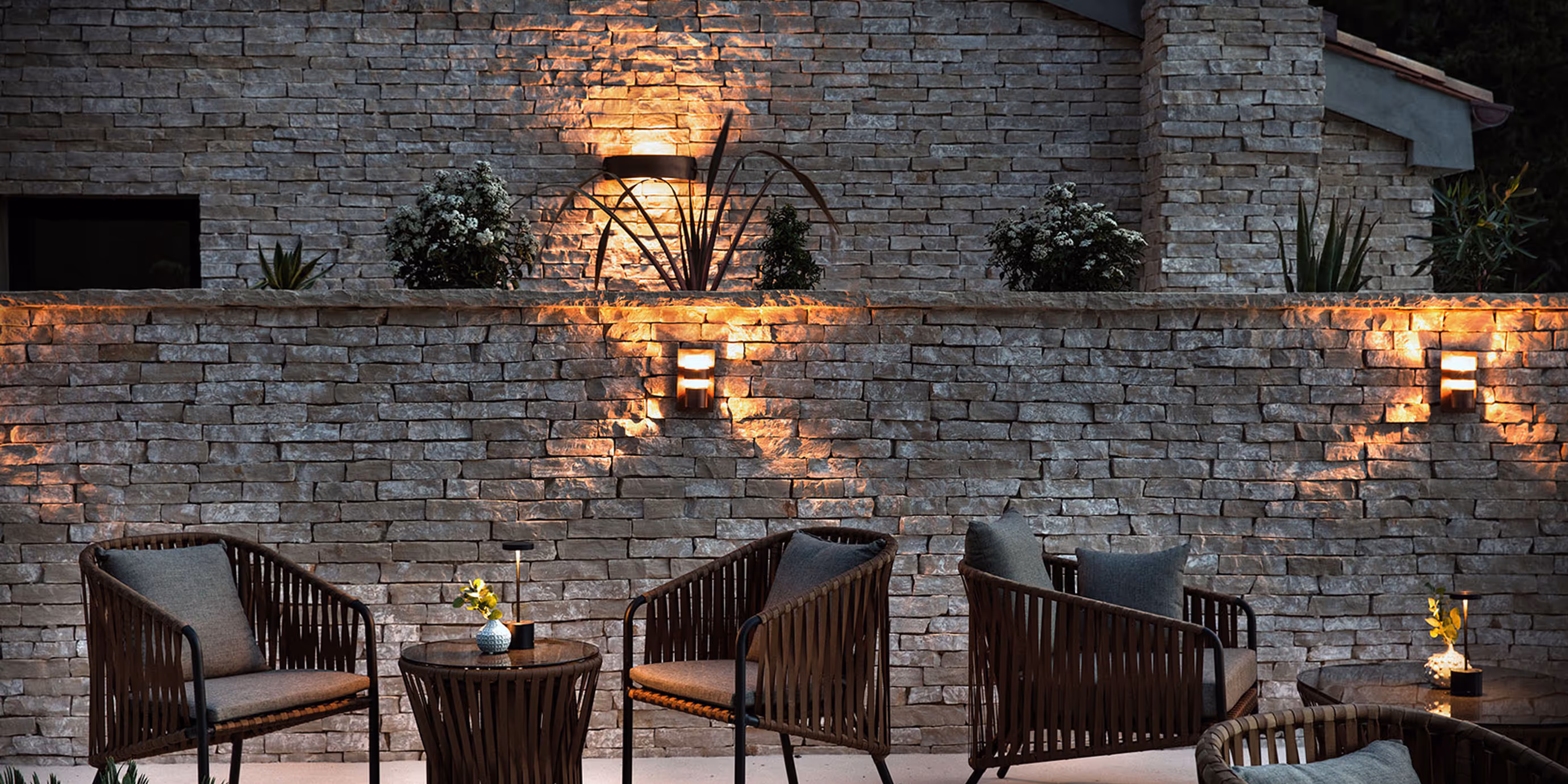 A terrace featuring natural looking chairs in front of warmly lit stone wall