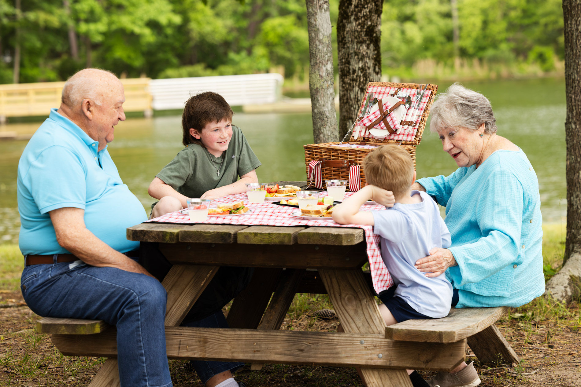 Two adults and two children having a picnic at a wooden table by a lake.