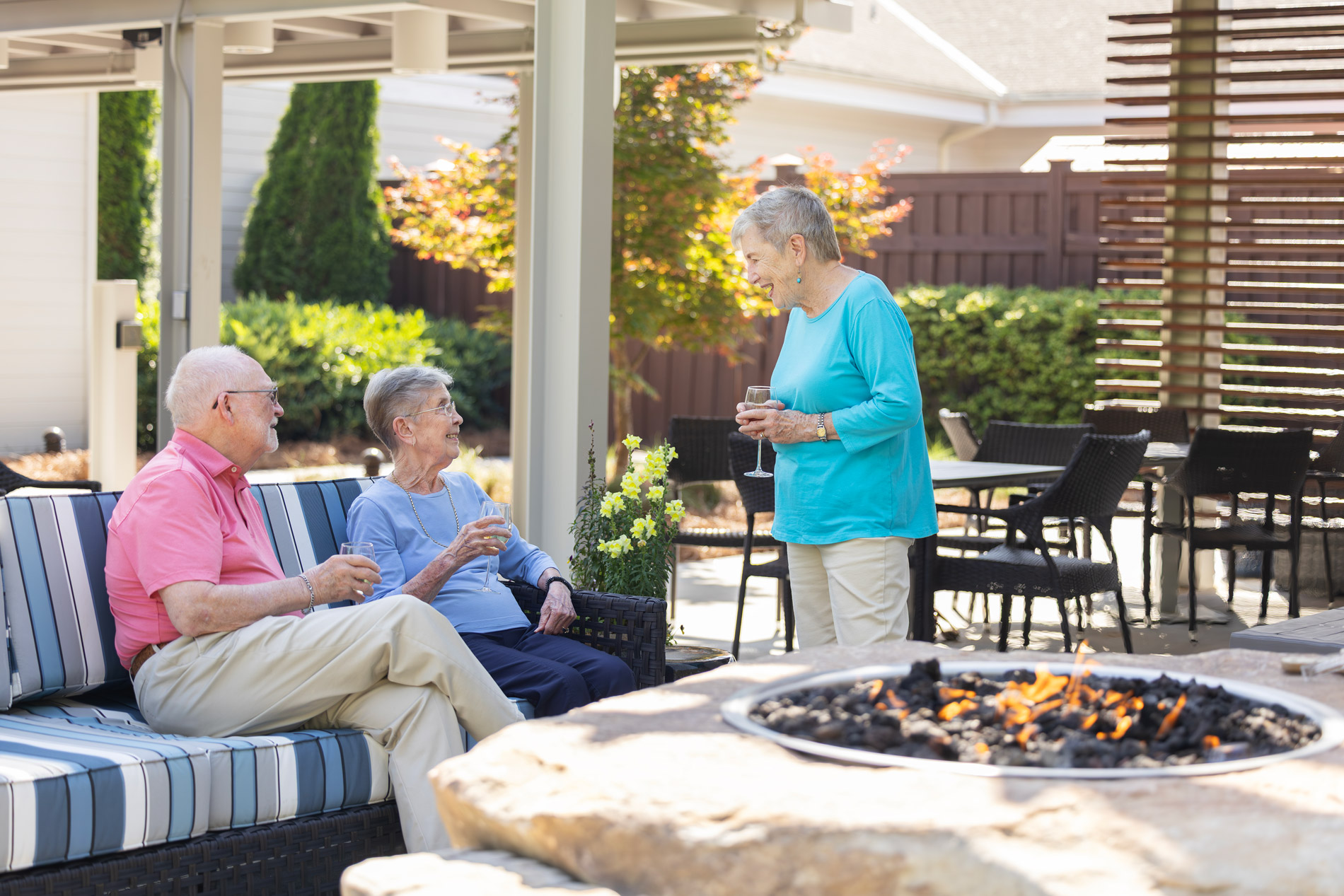 Three elderly people are talking and holding drinks near a fire pit in a patio area.