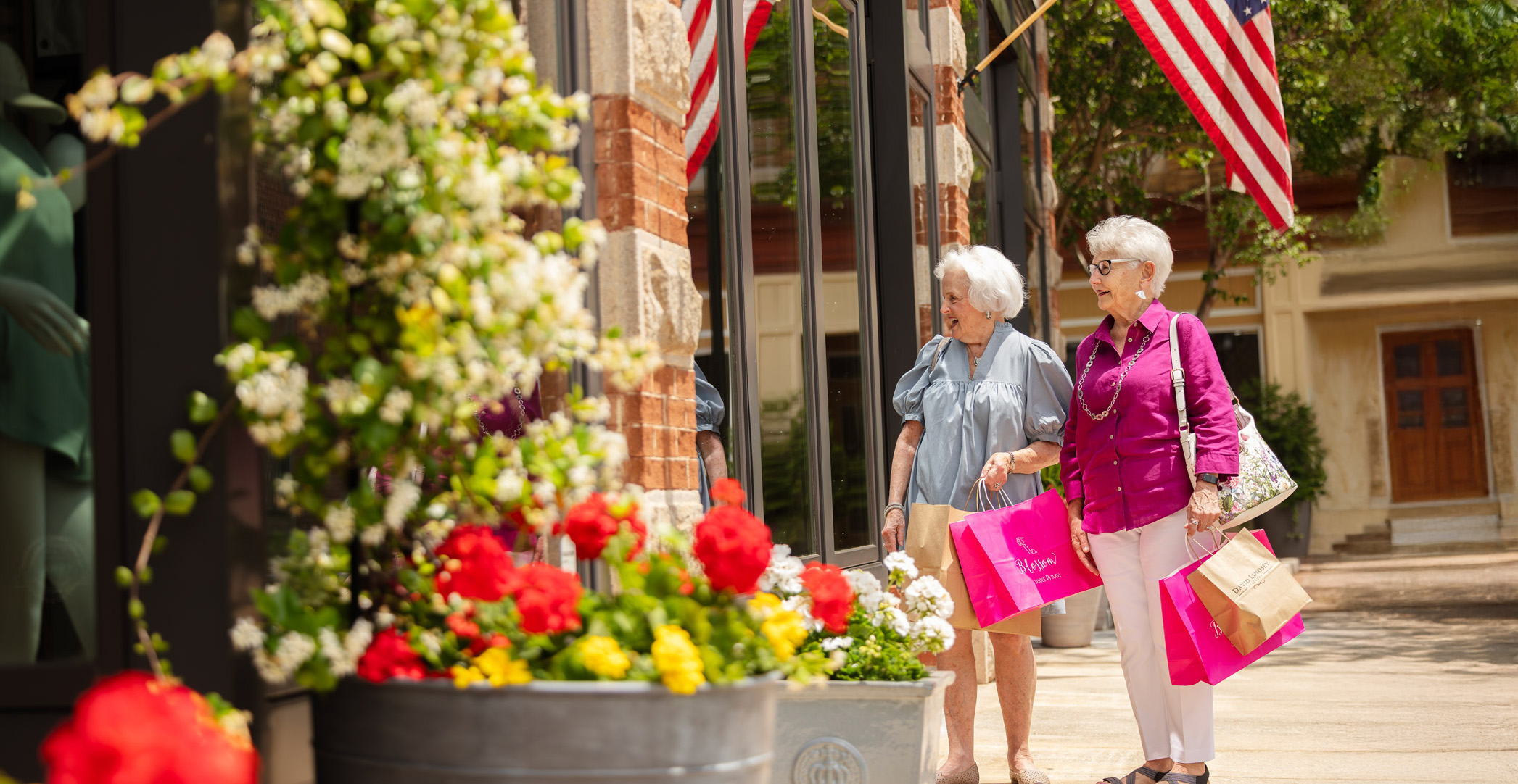 Two elderly women with shopping bags walk past a storefront with flowers and American flags.