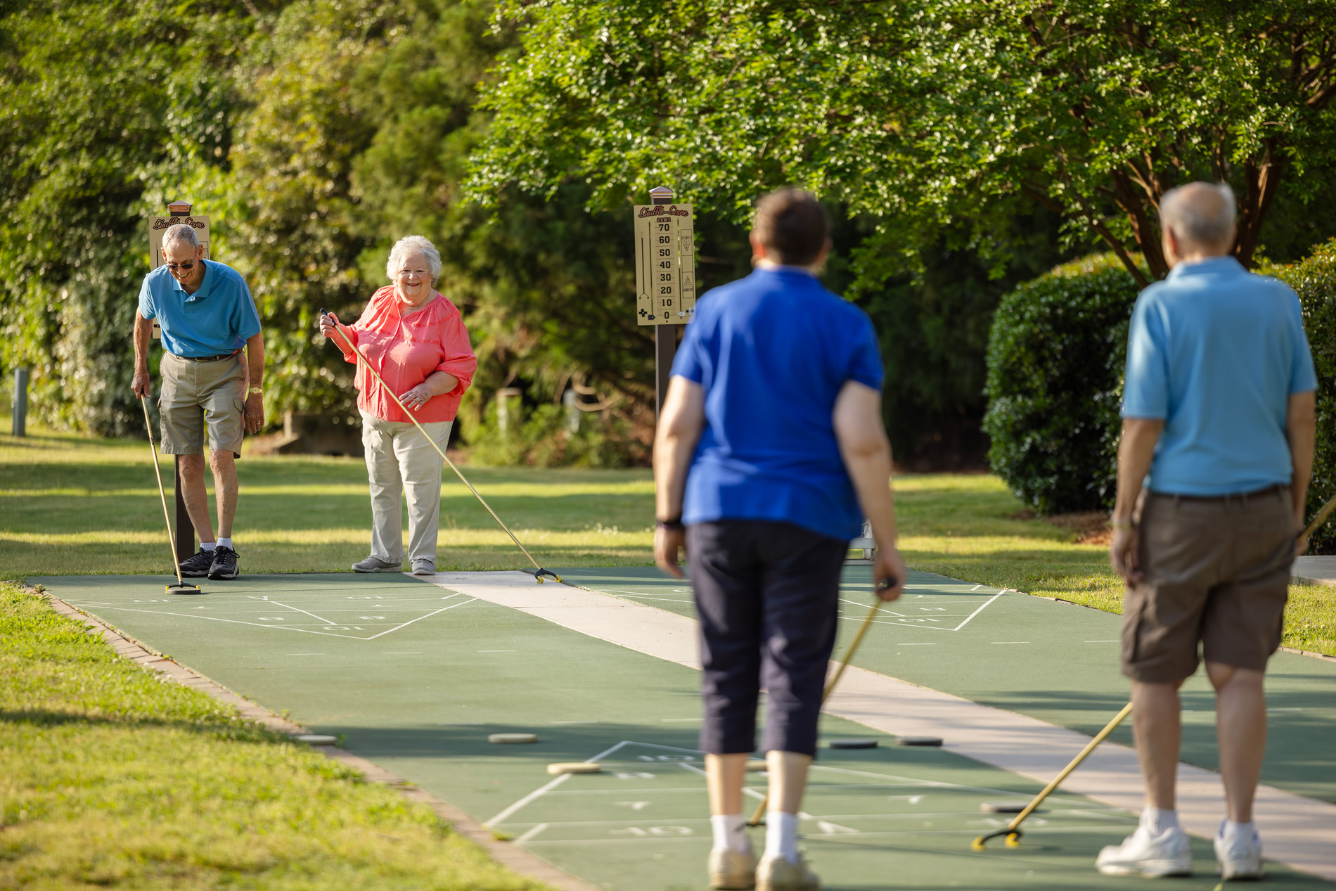 Four older adults are playing shuffleboard on an outdoor court in a park.