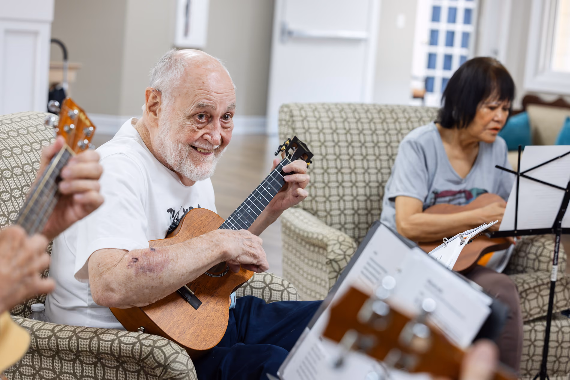 Elderly man smiles while playing a ukulele, surrounded by others playing instruments in a room.