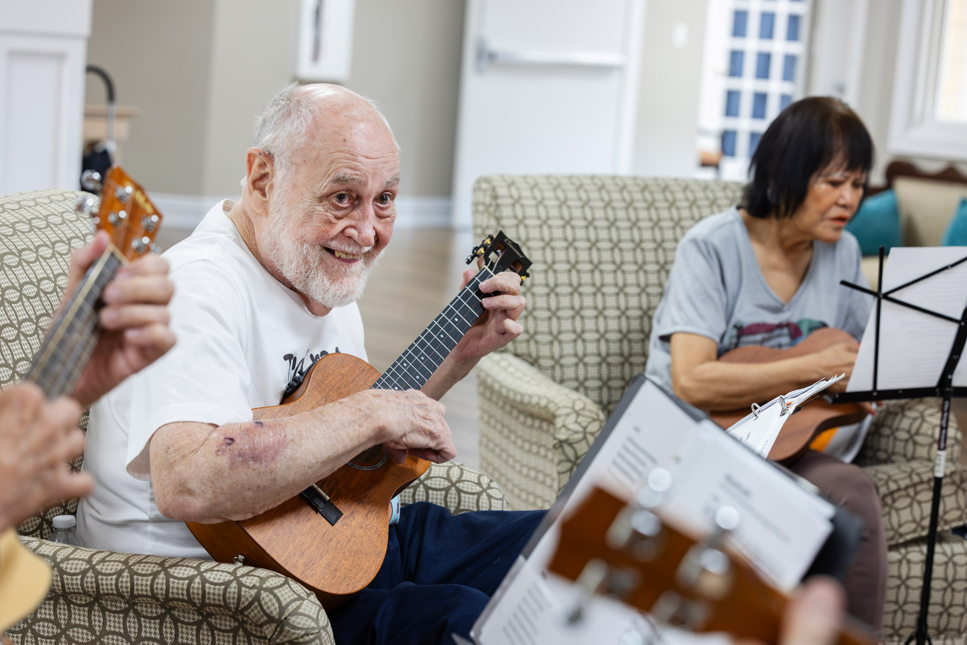 Elderly man smiles while playing a ukulele, surrounded by others playing instruments in a room.