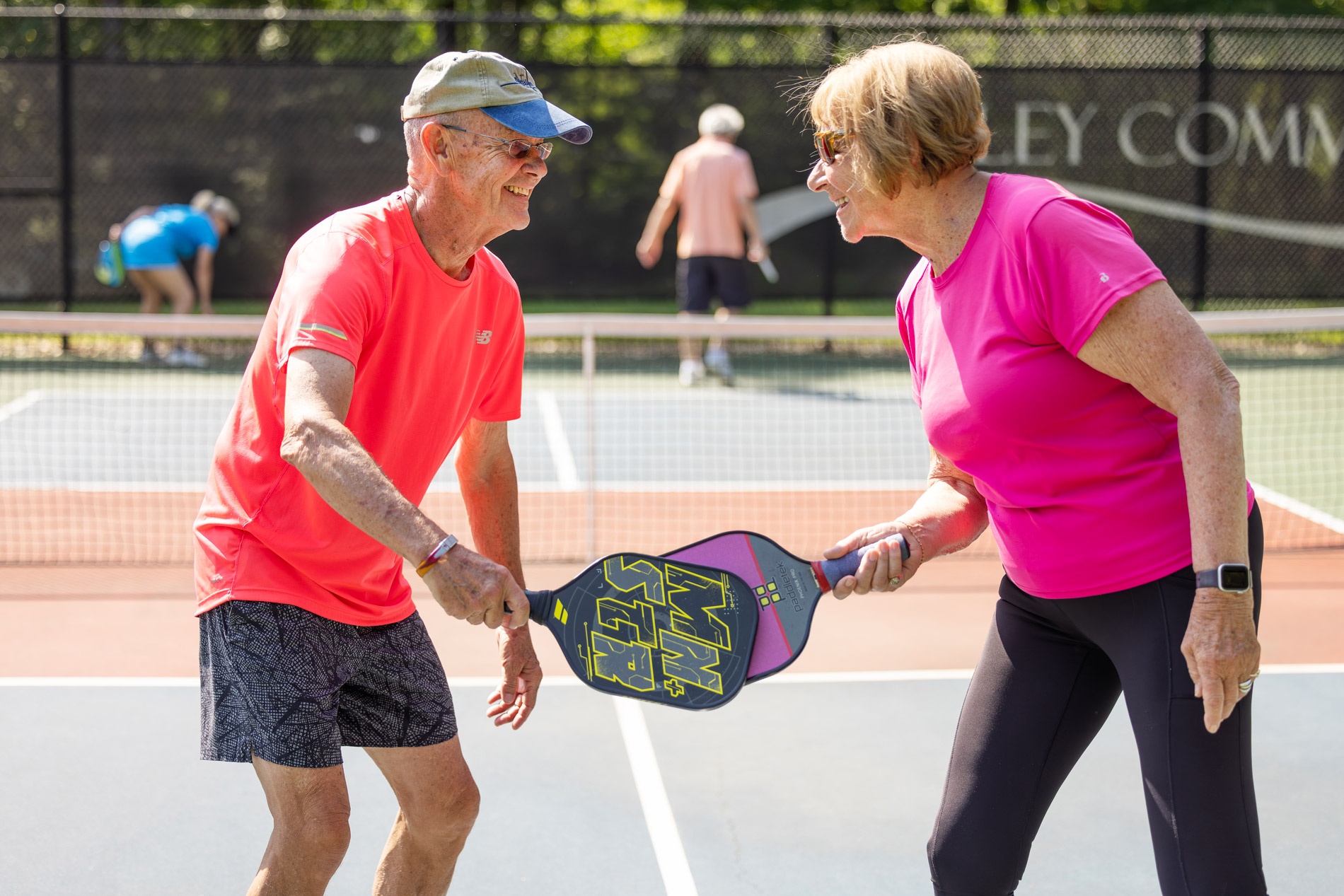 Two older adults smiling and holding pickleball paddles on a court.