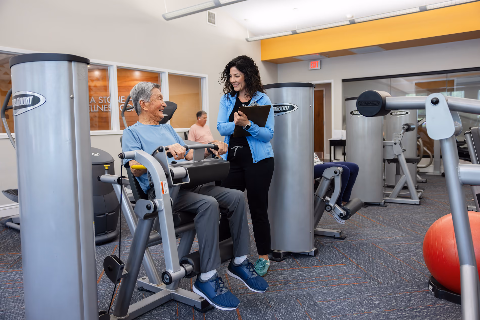 Older adult using exercise machine while a smiling woman with a clipboard stands nearby in a gym.