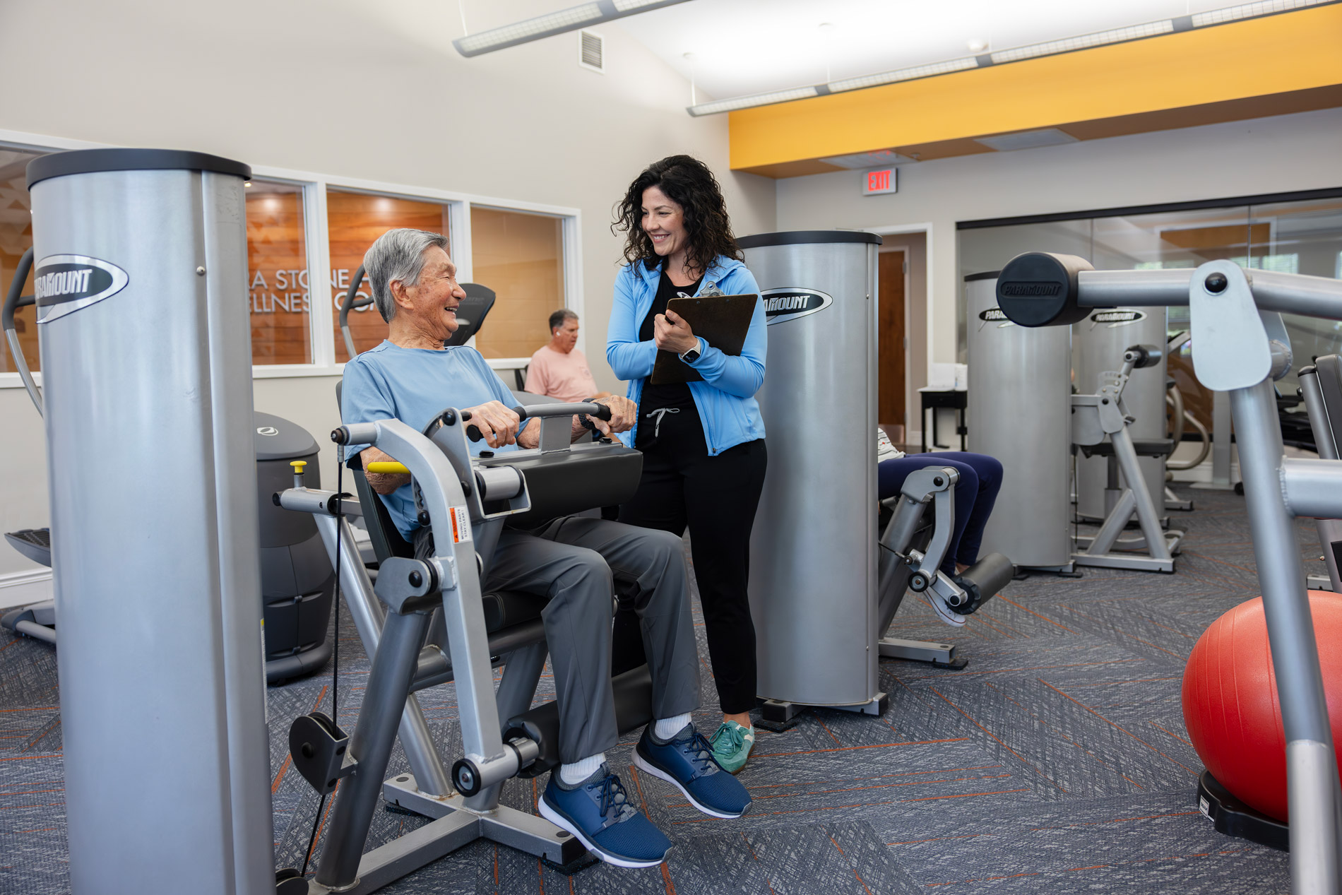 Older adult using exercise machine while a smiling woman with a clipboard stands nearby in a gym.