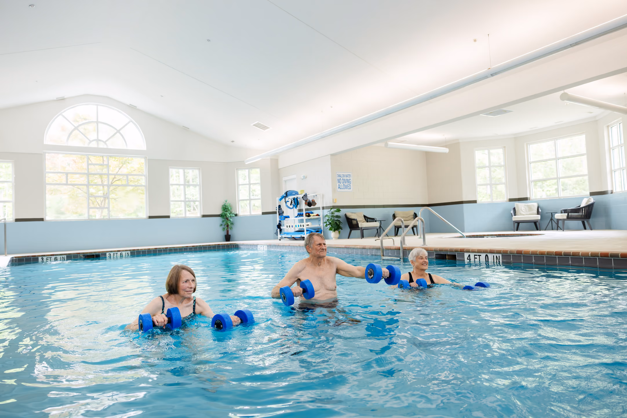 Three seniors doing water exercises with foam dumbbells in an indoor pool.