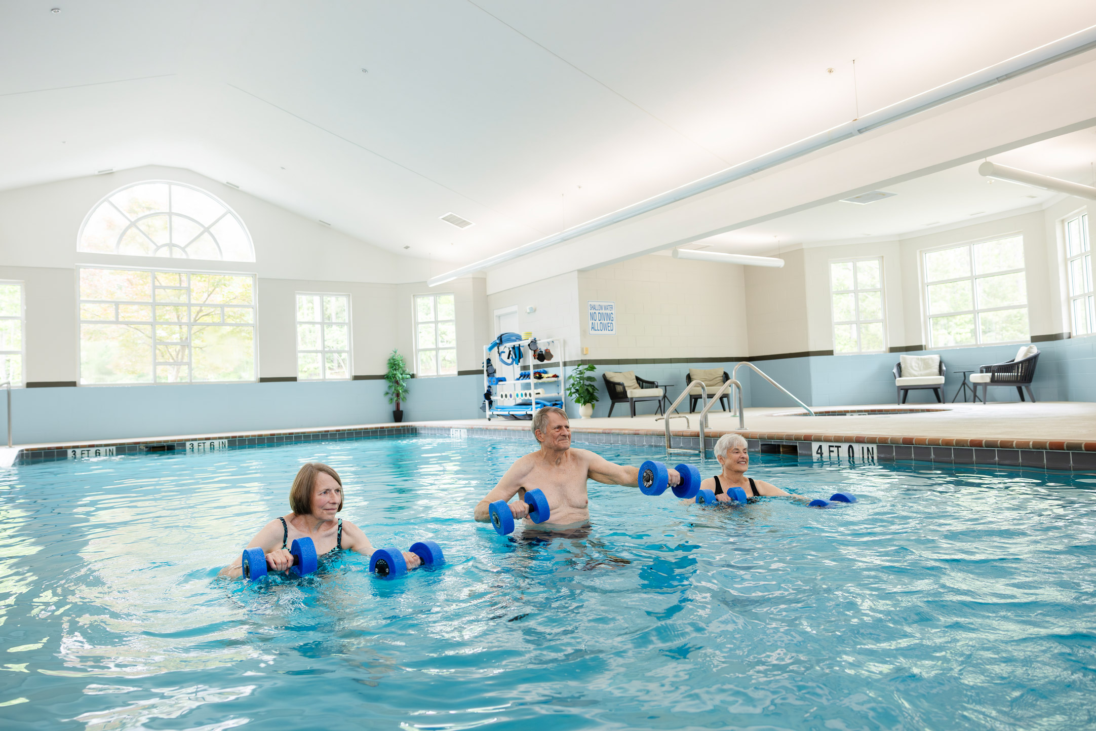 Three seniors doing water exercises with foam dumbbells in an indoor pool.