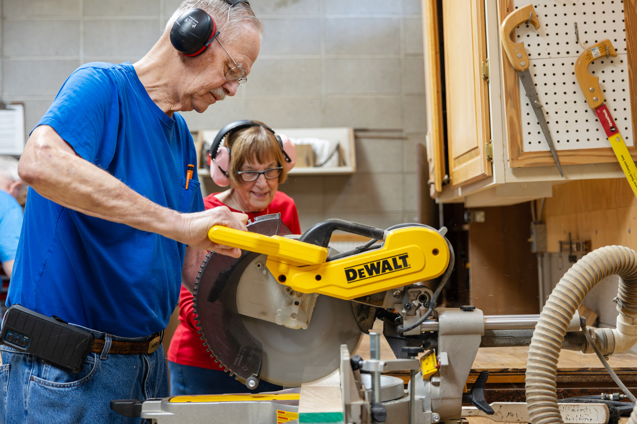 Man in blue shirt uses a yellow power saw while woman in red shirt watches closely.