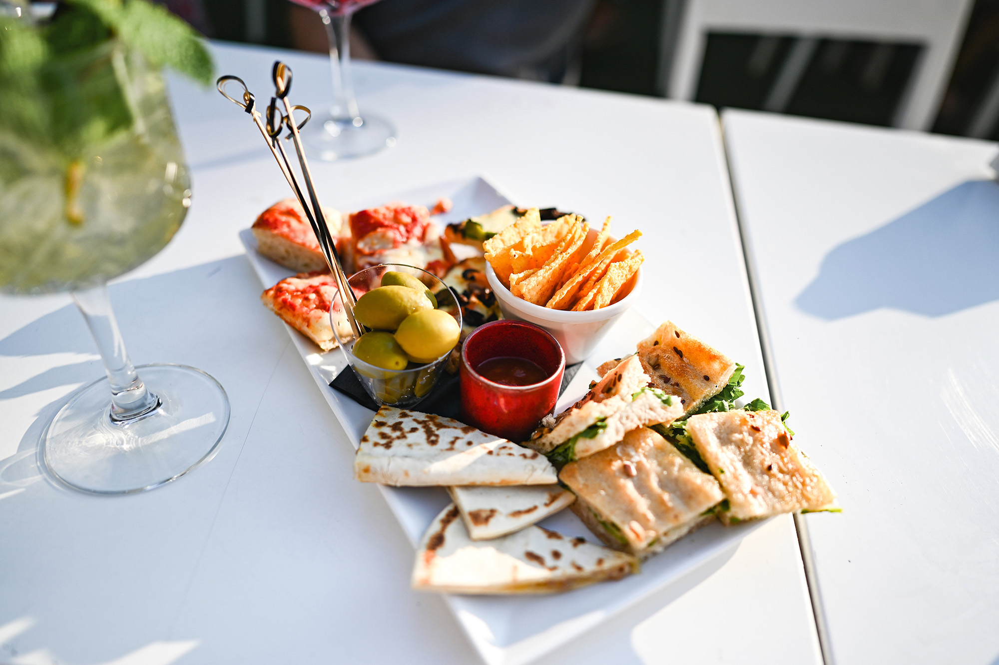 A white plate with assorted appetizers including sandwiches, olives, chips, and dipping sauce.