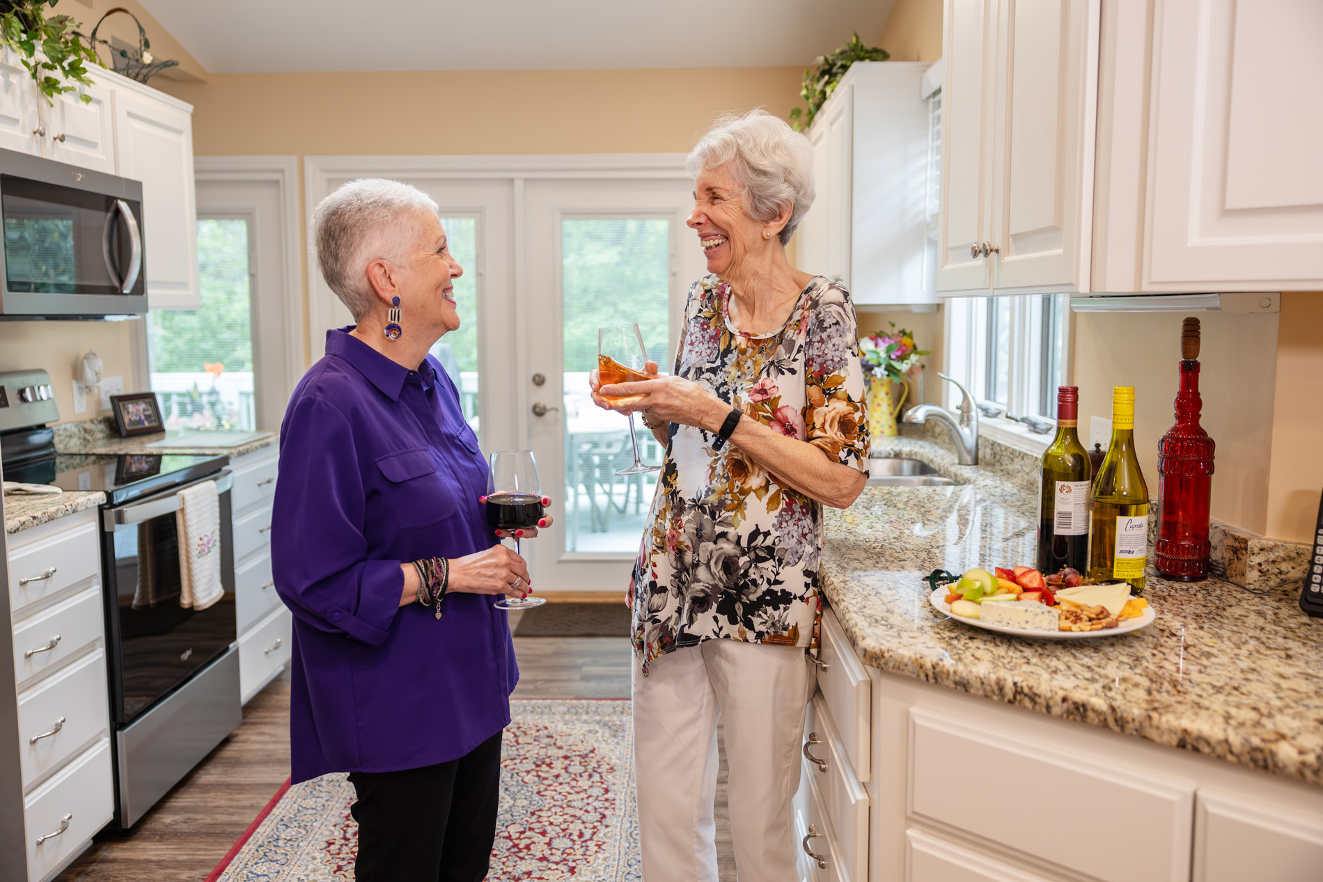 Two older women smiling and chatting in a kitchen, holding drinks and snacks.